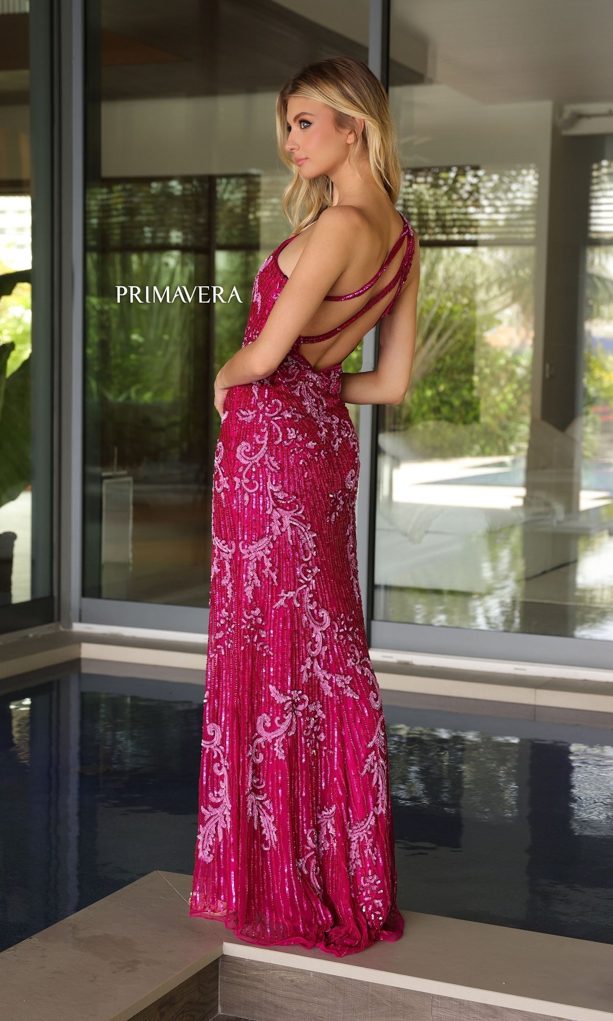 A woman stands indoors near a pool wearing the Primavera Strappy-Back Long Beaded Prom Dress 4124, a sleeveless, floor-length magenta gown with sequins and an open back.